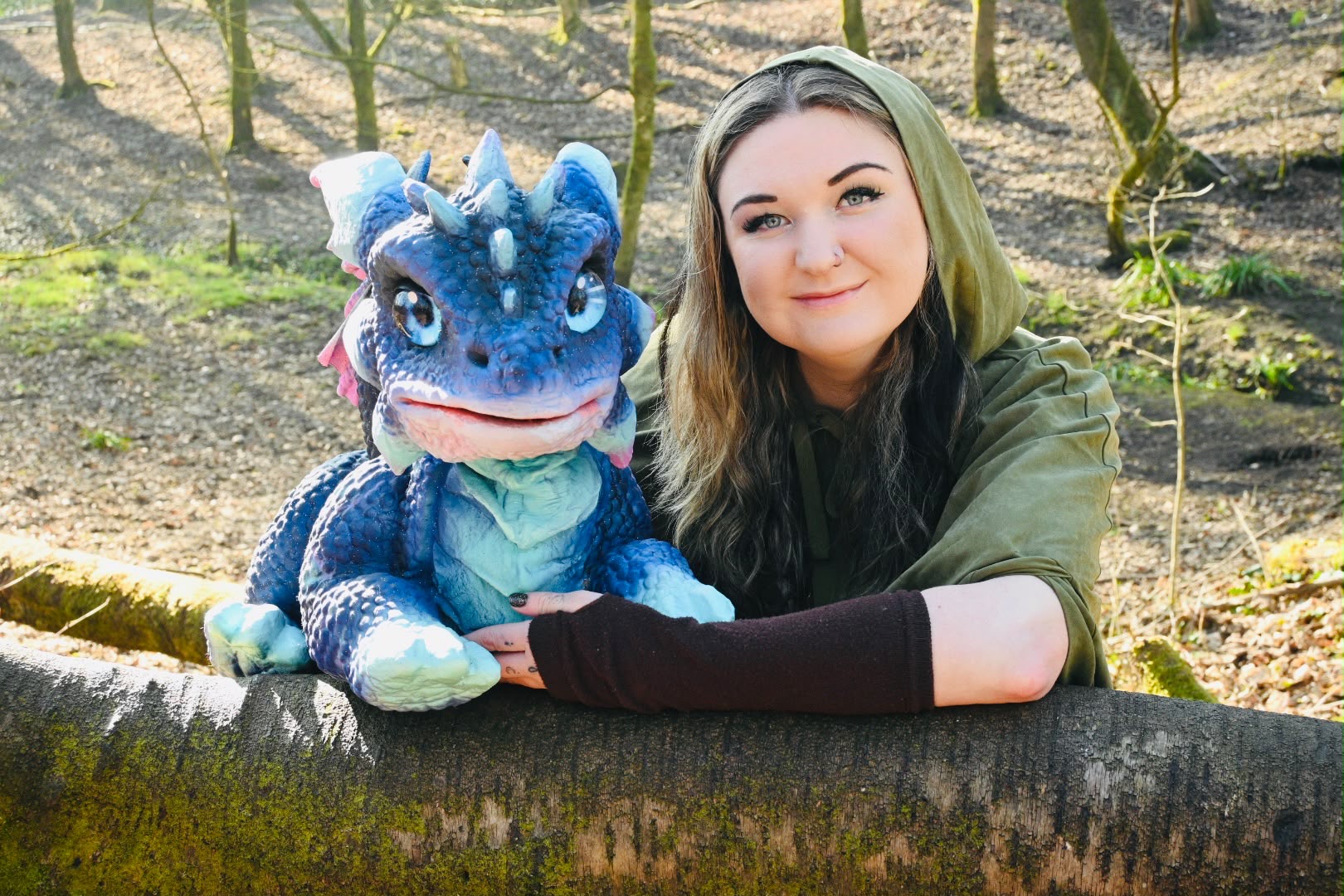 A cloaked handler with a blue baby dragon resting on a fallen tree in the woods