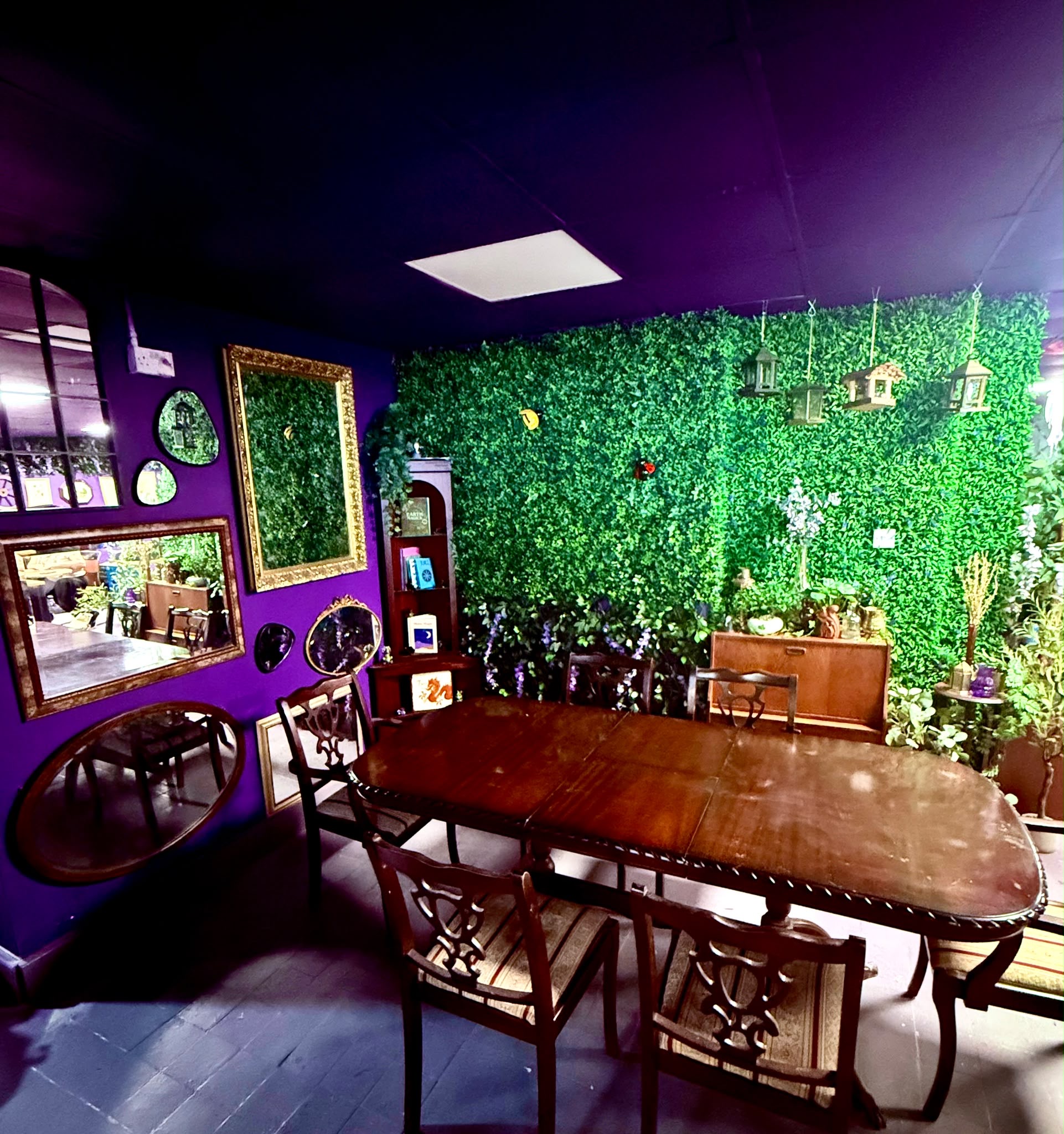 A wooden table and vintage chairs beside leafy walls and gold-framed mirrors