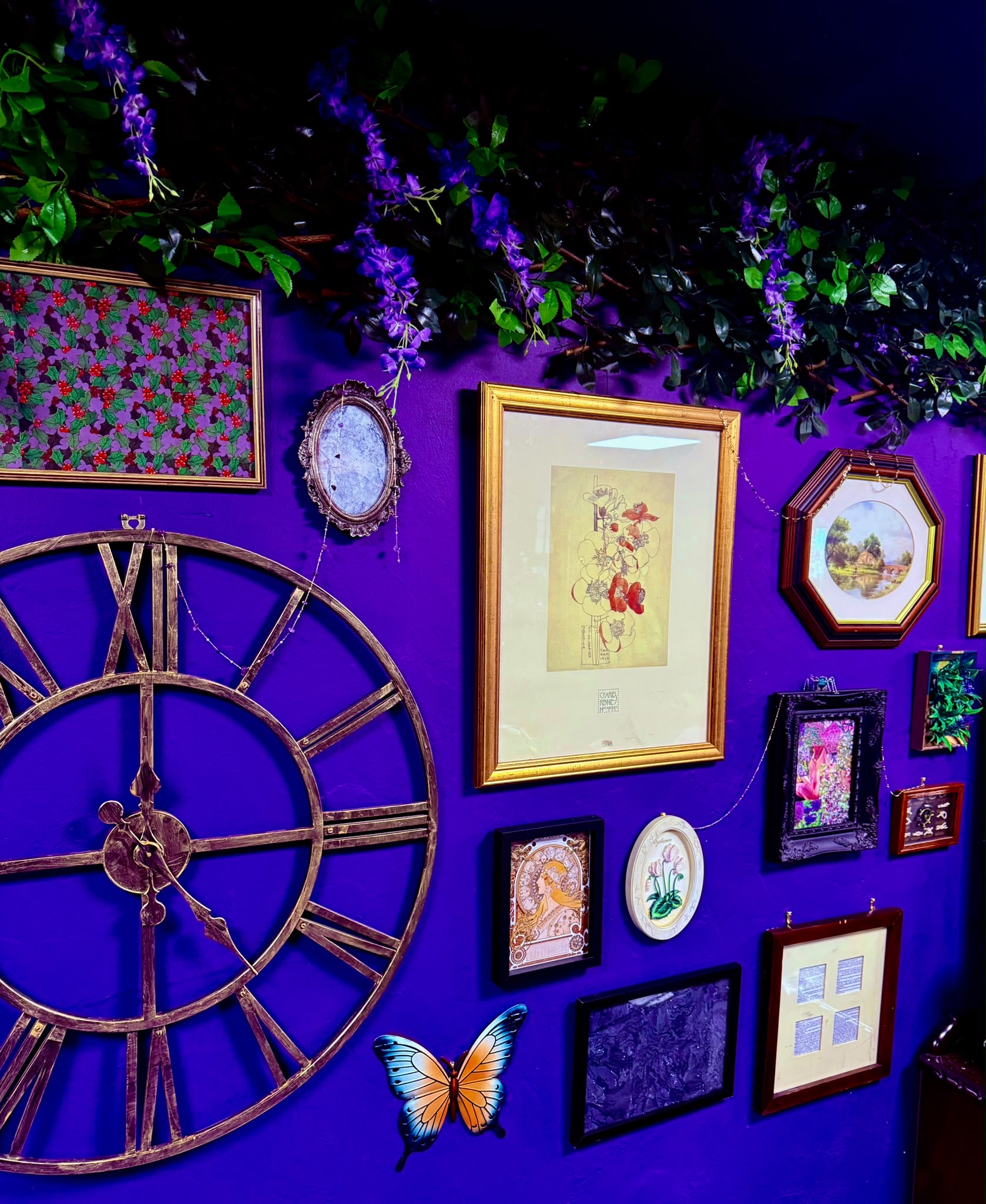 Gold picture frames and a large clock on a purple wall beneath wisteria