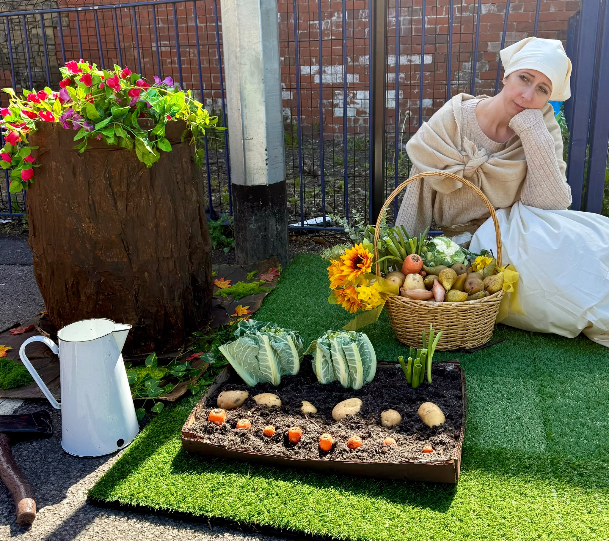 A costumed performer beside a garden stall with flowers and model vegetables
