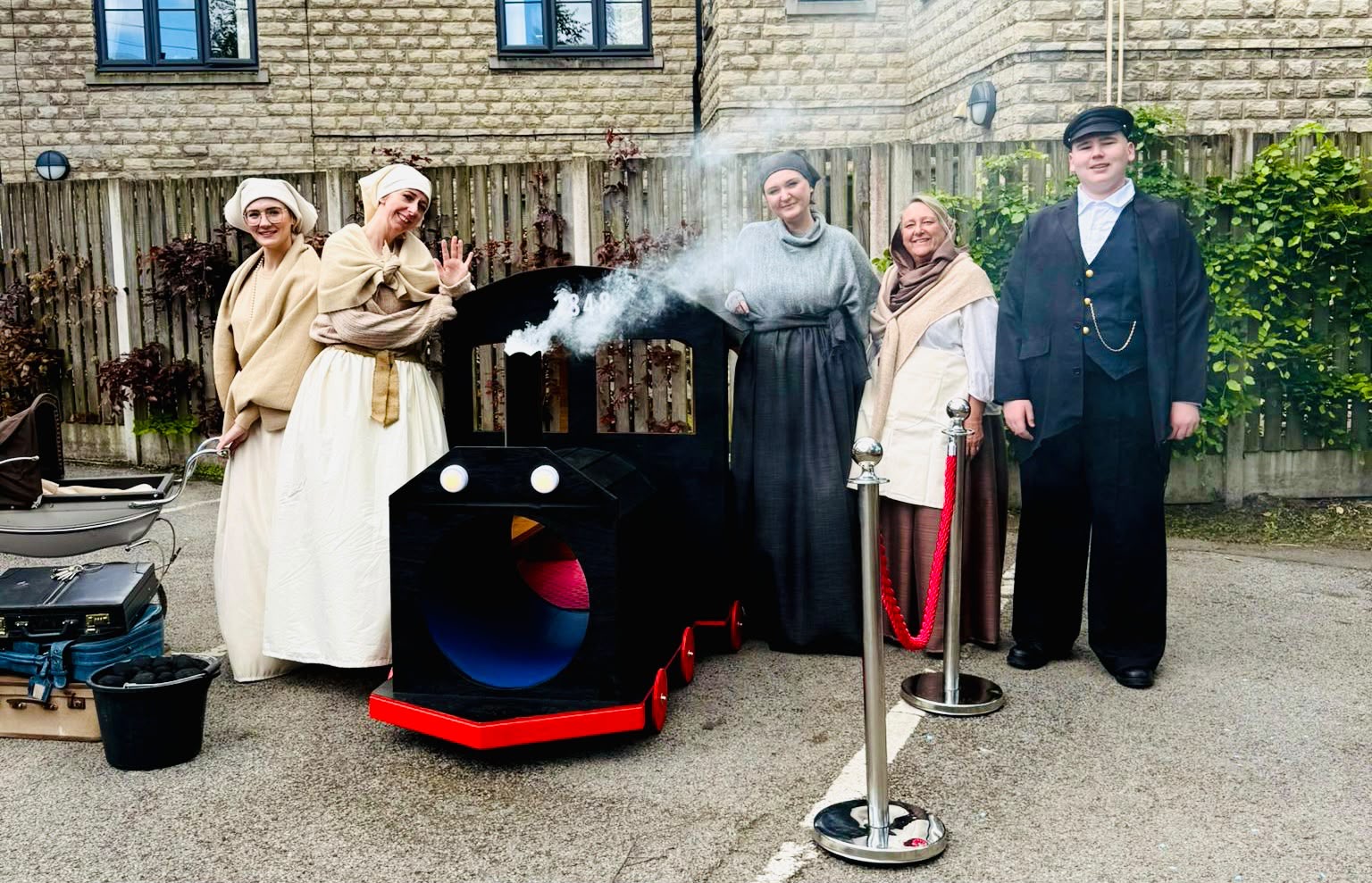 Five performers in period costumes beside a handmade steam train outdoors