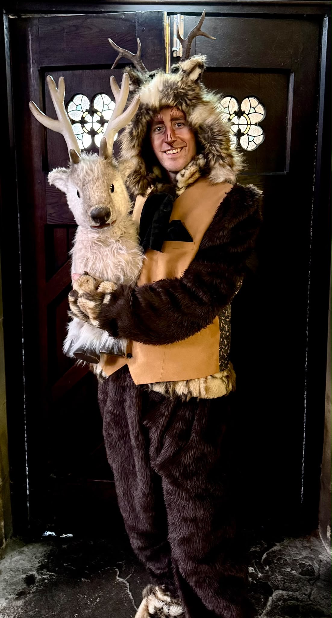 An antlered woodland character with a reindeer puppet by an ornate wooden door
